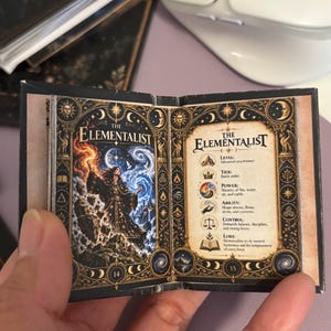 May include: A miniature open book titled The Elementalist, featuring a colorful illustration of a figure wielding fire and water on the left page and descriptive text about levels, tiers, powers, and lore on the right page, with ornate gold borders.