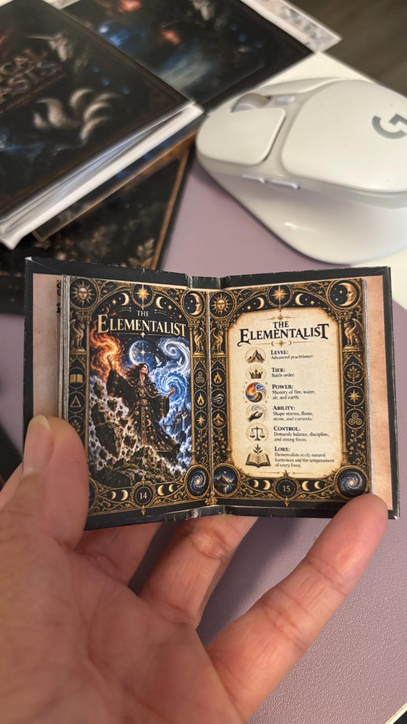 May include: A miniature open book titled The Elementalist, featuring a colorful illustration of a figure wielding fire and water on the left page and descriptive text about levels, tiers, powers, and lore on the right page, with ornate gold borders.