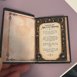May include: A miniature open book with black covers and cream-colored pages. The right page features a decorative border with celestial icons and the text: Opening the Arcane Orders. The text describes those who seek to shape mystery with will, study, and power.