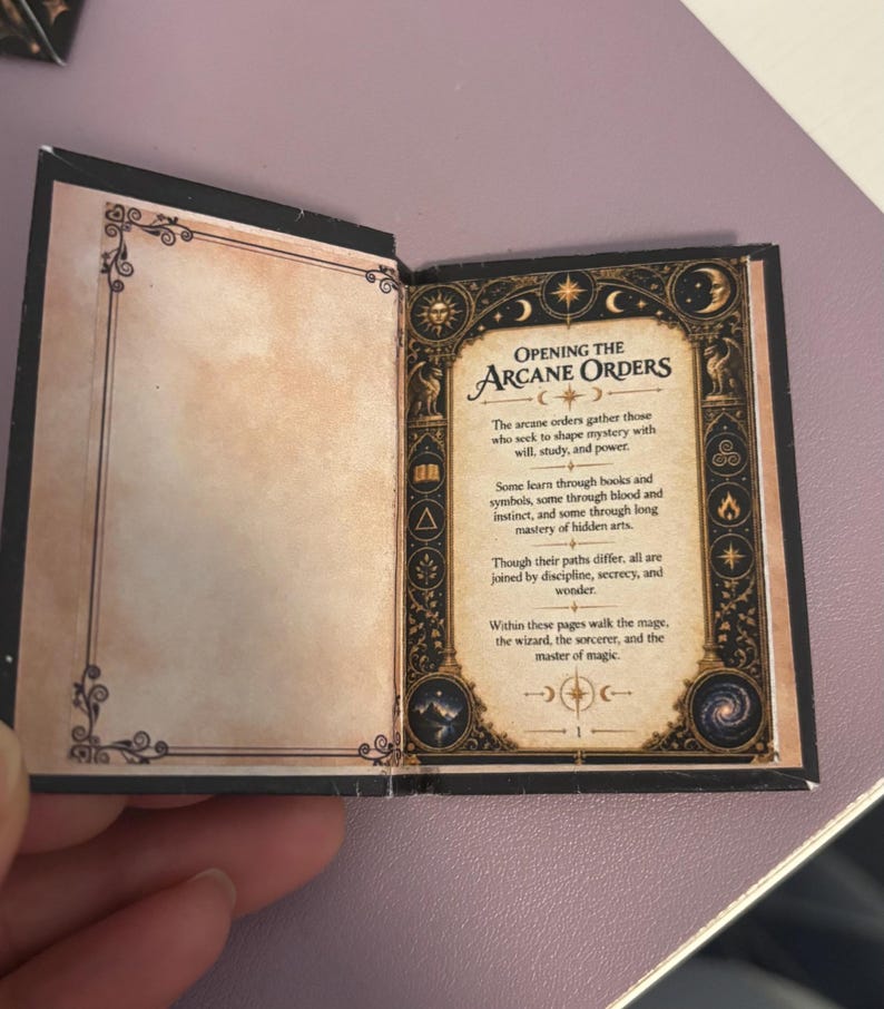 May include: A miniature open book with black covers and cream-colored pages. The right page features a decorative border with celestial icons and the text: Opening the Arcane Orders. The text describes those who seek to shape mystery with will, study, and power.