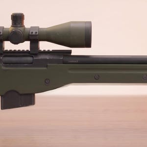May include: A bolt-action sniper rifle featuring an olive drab green thumbhole stock, a black barrel, and a mounted telescopic sight. The firearm includes a detachable box magazine, a black rubber butt pad, and a muzzle brake at the end of the barrel.