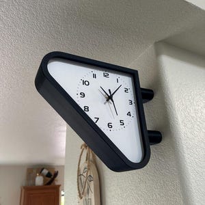 May include: A double-sided wall clock with a triangular black frame and a white face. The clock features black Arabic numerals from 1 to 12, black hour, minute, and second hands, and is mounted to a wall corner using two black cylindrical supports.