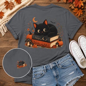 May include: A charcoal gray t-shirt featuring a graphic of a black cat resting on a stack of two books, surrounded by orange pumpkins, mushrooms, autumn leaves, a crescent moon, and a small bat. A smaller version of the same graphic is shown below.