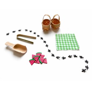 May include: A miniature picnic set featuring two woven baskets, a wooden scoop, wooden tongs, a green and white checkered cloth, several small pink and green watermelon slices, and a trail of small black plastic ants arranged in a curved line.