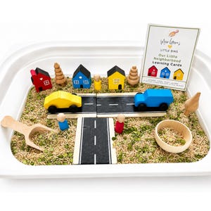 May include: A sensory play bin containing green split peas and rice, featuring wooden toy houses in red, blue, and yellow, two wooden cars, road segments, wooden peg people, a wooden scoop, a small wooden bowl, and a card labeled Miss Lossa's Little Bins.