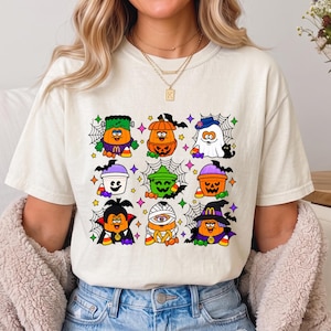 May include: A cream-colored t-shirt featuring a graphic print of nine cartoon characters dressed in Halloween costumes, including a pumpkin, ghost, vampire, and mummy, accented with spider webs, candy corn, and colorful star shapes.