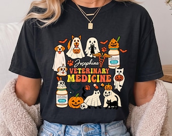 May include: A black t-shirt featuring a colorful Halloween-themed graphic with the text Josephine VETERINARY MEDICINE. The design includes ghosts, cats, dogs, pumpkins, medicine bottles, a stethoscope, and a caduceus symbol in orange and yellow.