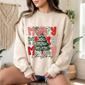 Op de afbeelding: Een beige sweater met ronde hals en een centrale afbeelding van een groene kerstboom met een rood-witte strik in ruitpatroon. Het ontwerp bevat rood en groen gedessineerde letters die Merry Christmas spellen in een opvallend lettertype.