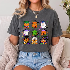 May include: A charcoal gray short-sleeve t-shirt featuring a graphic print of nine cartoon characters dressed in various Halloween costumes, including a pumpkin, ghost, vampire, and mummy, accented with spiderwebs and colorful candy corn.