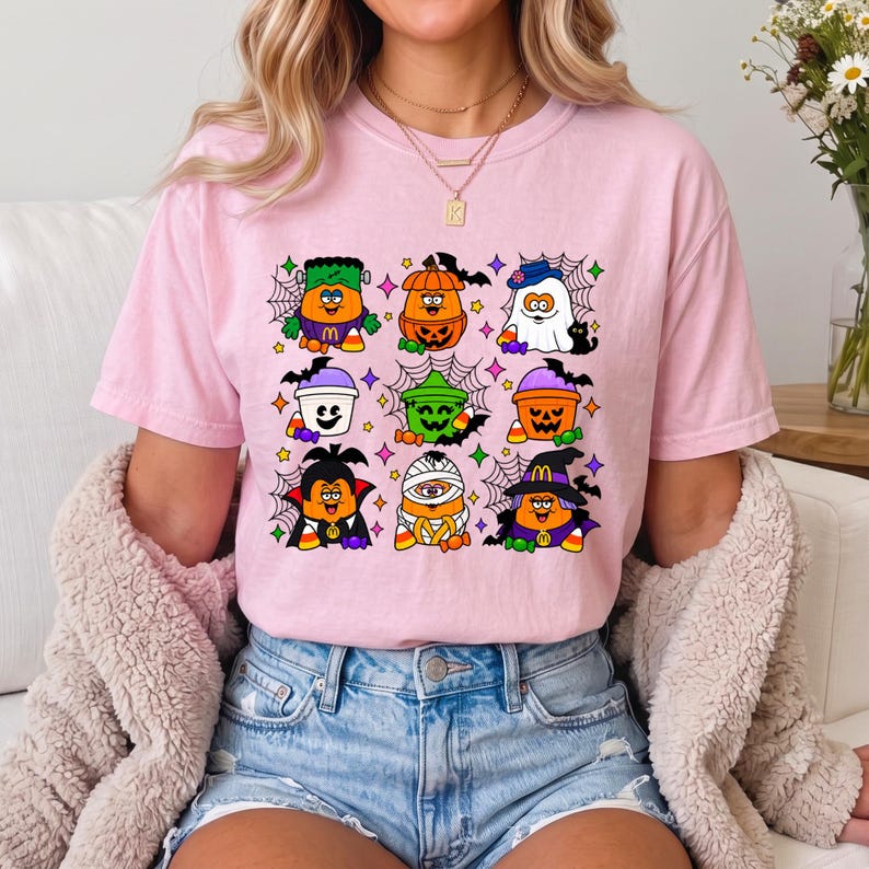 May include: A light pink t-shirt featuring a grid of nine colorful Halloween-themed illustrations of orange characters dressed as various figures including a vampire, a mummy, a ghost, and a pumpkin, accented with spiderwebs and small stars.