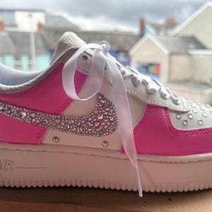 May include: A white and hot pink sneaker featuring a side swoosh logo covered in clear crystals. The shoe includes white ribbon laces, a thick white rubber sole with the text AIR embossed on the side, and small crystal embellishments on the toe box.