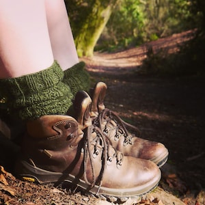 May include: A pair of brown leather hiking boots with dark brown laces and Vibram soles, worn with thick, forest green knitted wool socks that feature a ribbed texture and a folded cuff design, covering the lower legs above the ankle.