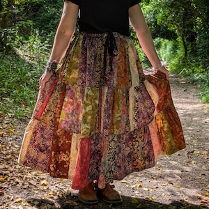 Puede incluir: Una falda larga de varios niveles con un diseño de patchwork de diversas telas en tonos marrón, óxido, verde oliva y crema. La falda tiene una cintura fruncida con un cordón negro y se combina con una camiseta negra lisa de manga corta.