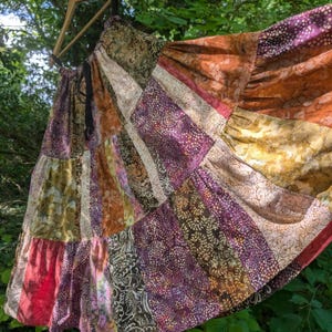 Op de afbeelding: Een lange, gelaagde rok met een patchworkontwerp van diverse stoffen in tinten paars, oranje, bruin en goud. De rok is opgebouwd uit verticale panelen met uiteenlopende bloem- en geometrische patronen en is voorzien van een zwart trekkoord.