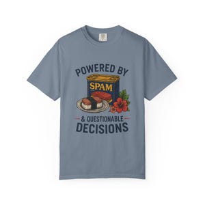 Op de afbeelding: Een leiblauw t-shirt met korte mouwen en een afbeelding van een blauw Spam-blik, een stuk Spam-musubi op een bord en een rode hibiscusbloem. De tekst luidt POWERED BY SPAM & QUESTIONABLE DECISIONS in donkerblauwe hoofdletters.