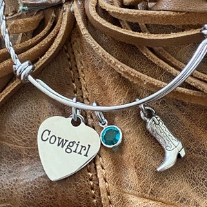 May include: A silver bangle bracelet with a heart charm that says "Cowgirl", a turquoise gemstone charm, and a silver boot charm.