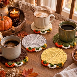 May include: A set of five round crochet coasters in cream, light grey, dark brown, olive green, and yellow. Each coaster features a decorative border of small, colourful crocheted flowers in orange, white, and green along the bottom edge.
