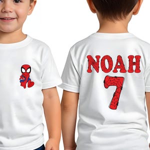 May include: A white short-sleeved t-shirt featuring a small Spider-Man graphic on the front left chest. The back of the shirt displays the name NOAH in red spiderweb-patterned letters above a large red spiderweb-patterned number 7.