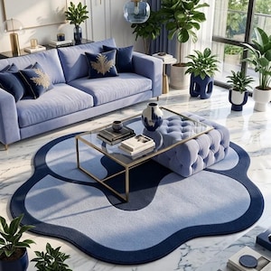 May include: A large, organic-shaped area rug featuring a layered design in shades of navy and light blue. The rug has a smooth, solid-colored border and a soft, textured center, creating a distinct contrast against the white marble floor.