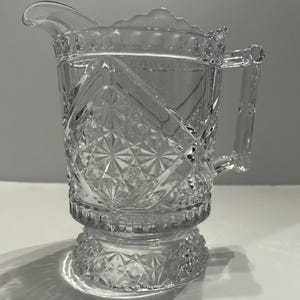 May include: A clear glass pitcher featuring an intricate pressed pattern with diamond and starburst motifs. The vessel has a scalloped rim, a sturdy handle, and a wide circular base, all decorated with a repeating geometric texture throughout.