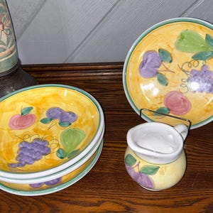 May include: A set of three ceramic plates and a small pitcher featuring a hand-painted yellow background with green, purple, and pink fruit motifs including grapes, pears, and peaches. The plates have a green rim, and the pitcher has a white interior.