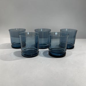 May include: A set of five matching drinking glasses made of translucent blue glass. Each glass features a smooth, cylindrical body with a slightly flared rim and a thick, weighted base that provides stability and a distinct profile for each vessel.