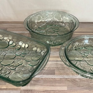 May include: A set of three clear, light green glass baking dishes featuring a textured fruit pattern on the exterior. The collection includes one rectangular dish, one round bowl, and one round shallow dish, all with a consistent embossed fruit design.