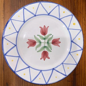 May include: A round white ceramic plate featuring a central hand-painted design of four red tulips with green stems and leaves arranged in a cross pattern. The rim is decorated with a blue triangular geometric border and small yellow dots.