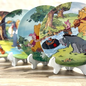 May include: Four round ceramic decorative plates displayed on white stands. Each plate features colorful illustrations of Winnie the Pooh, Piglet, Eeyore, and Roo in a forest setting, depicting scenes with a stream, trees, and grassy landscapes.