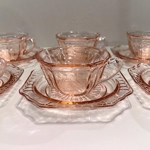 May include: A set of pink depression glass teacups and matching saucers. Each piece features a translucent light pink hue and a molded floral pattern with scalloped edges. The cups have a curved handle and the saucers have a central circular base.