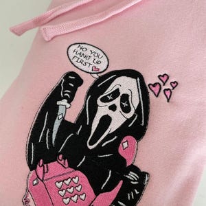 Könnte beinhalten: Ein hellrosa Kapuzenpullover mit einer Stickerei einer schwarz gekleideten Figur, die ein Messer und ein rosa Wählscheibentelefon hält. Eine Sprechblase über der Figur zeigt den Text "NO YOU HANG UP FIRST" mit einem kleinen rosa Herz.