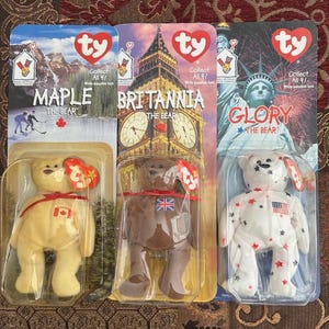 May include: Three Ty Beanie Babies in their original packaging. The bears are named Maple, Britannia, and Glory. Maple is tan with a Canadian flag. Britannia is brown with a Union Jack. Glory is white with red and blue stars.