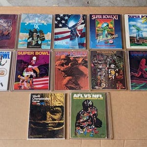May include: A collection of vintage Super Bowl and AFL vs NFL game programs. The covers feature colorful illustrations of football players, the American flag, and the Super Bowl trophy. The programs are encased in clear protective sleeves.