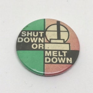 May include: A round button with a green, black, and pink background. The button has the text "SHUT DOWN OR MELT DOWN" in black letters.