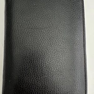 May include: A rectangular black notebook with a textured, pebbled surface. The cover is solid black and features stitched edges along the perimeter, providing a clean and uniform appearance for this functional stationery item used for writing.