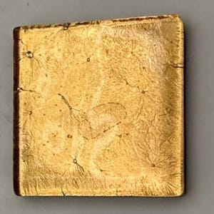 May include: A square, gold-colored decorative tile with a textured surface. The tile has a network of small, dark spots and lines across its surface, creating a unique pattern. The edges of the tile are a darker shade of gold.