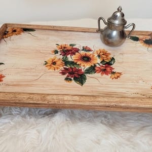 May include: A rectangular wooden breakfast tray with folding legs, featuring a light wood grain finish and a central floral motif of sunflowers and red blossoms. A small, metallic, two-handled lidded vessel sits on the tray surface.