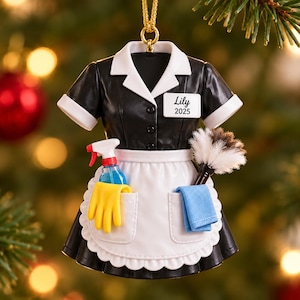 以下が含まれることがあります： Lily 2025と書かれた名札が付いた、黒と白のメイド服のオーナメント。白いエプロンには2つのポケットがあり、青いスプレーボトル、黄色いゴム手袋、羽はたき、青い布が入っています。