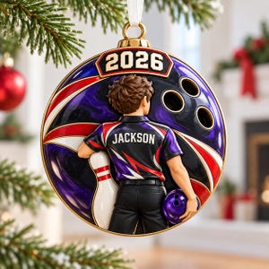 May include: A round Christmas ornament featuring a bowling theme with the year 2026 at the top. A figure in a black, purple, and red shirt with the name JACKSON printed on the back holds a bowling ball and stands next to a bowling pin.