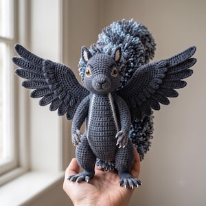 May include: A dark gray crocheted squirrel figurine featuring large, spread-out wings and a voluminous, textured tail in shades of blue and gray. The creature has amber-colored eyes, small white claws on its feet, and a white stripe down its chest.
