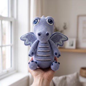 May include: A light purple crocheted dragon plush toy featuring large dark blue and white eyes, detailed wings with intricate stitching, and horizontal stripes on the torso. The creature has a rounded snout, small arms, and feet with dark blue accents.