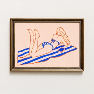 Puede incluir: Una lámina de arte lineal enmarcada que muestra a una persona de pelo largo tumbada boca abajo sobre una toalla de rayas azules y blancas. La persona lleva una braguita de bikini de rayas azules y blancas y un top azul. Un pequeño libro azul descansa sobre la toalla.