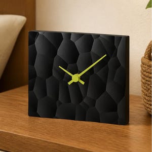 May include: A rectangular table clock with a matte black face featuring a three-dimensional geometric pattern of irregular polygons. The clock has bright lime green hour and minute hands centered on the dark, textured surface.