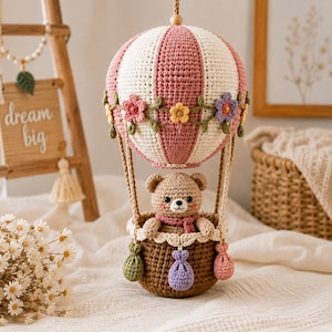 Könnte beinhalten: Ein gehäkeltes Heißluftballon-Ornament mit einem rosa-weiß gestreiften Ballon, der mit kleinen bunten Blumen verziert ist. Ein kleiner brauner Häkelbär sitzt im dunkelbraunen Korb, der mit drei kleinen hängenden Beuteln geschmückt ist.