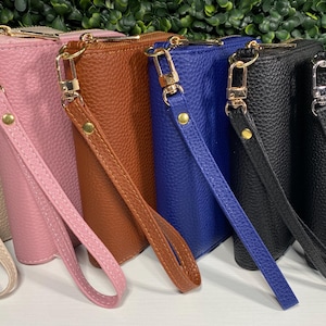 May include: A row of six rectangular wristlet wallets with a pebbled texture, each featuring a gold-tone zipper, a matching detachable wrist strap with a gold-tone clasp, and a gold-tone rivet. The wallets are beige, pink, brown, blue, and two black.