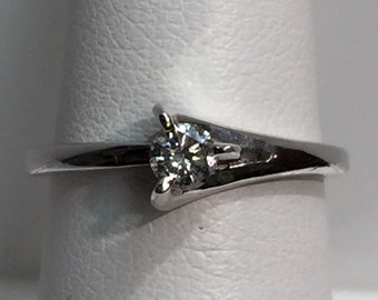 Könnte beinhalten: Ein silberner Ring mit einem einzelnen runden Diamanten, der in einer Krappenfassung sitzt. Die Band ist dünn und hat ein geschwungenes Design.