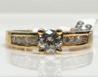 May include: A gold ring featuring a large, round, gray diamond center stone. The band has a channel setting with smaller diamonds on each side. The ring is displayed on a white surface, reflecting the ring's design.