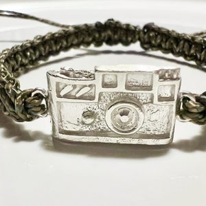 May include: A green braided bracelet with a silver camera charm. The camera charm has a detailed design with a lens and a viewfinder.