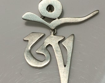 May include: A silver pendant featuring the Om symbol, a sacred sound in Hinduism and Buddhism. The pendant is made of metal and has a smooth, polished finish.