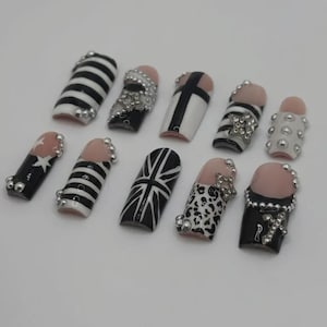 May include: A set of ten press-on nails featuring a variety of black, white, and nude designs. Patterns include horizontal stripes, a Union Jack, leopard print, and solid black tips. Embellishments include silver studs, rhinestones, and cross charms.